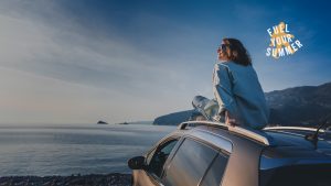 A woman wearing sunglasses, sitting on top of her car and looking out at the water. nomnom "Fuel Your Summer" logo is in the upper right corner.